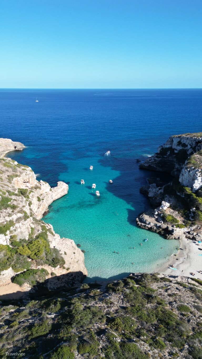 Hochformat-Luftaufnahme der Cala Màrmols mit Strand, Felswänden und Booten vor dem offenen Meer