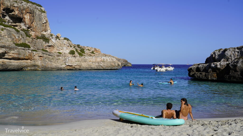 Heller Strand und türkisblaues Wasser der Cala Màrmols mit Badegästen und einem Paddleboard