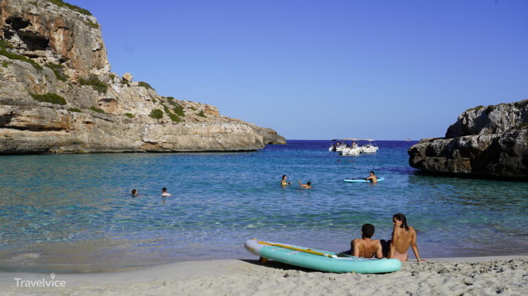 Heller Strand und türkisblaues Wasser der Cala Màrmols mit Badegästen und einem Paddleboard